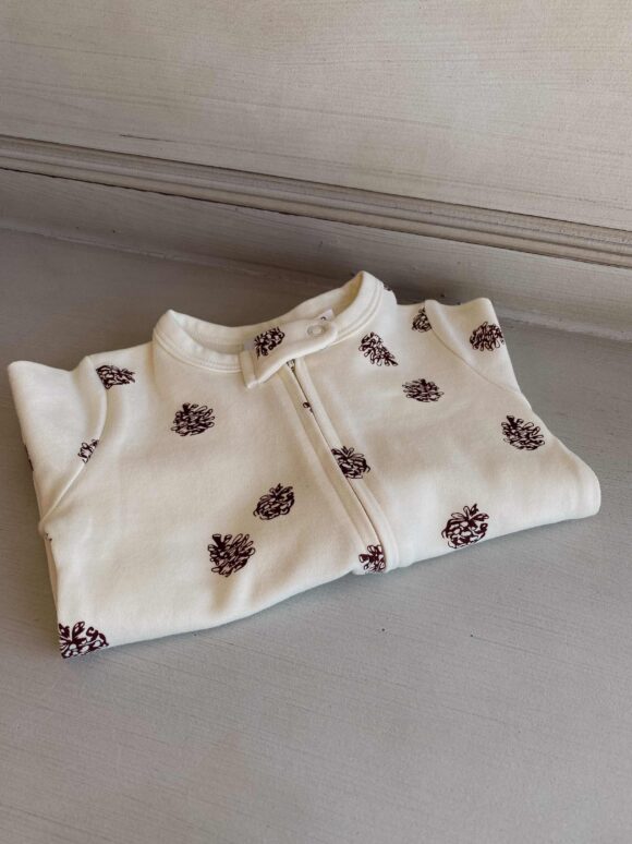 Baby long sleeved romper with zip front in light beige and acorn design is folded up and displayed on top of a piece of furniture.