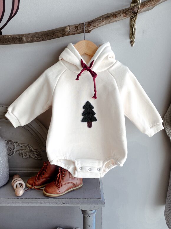 thick Christmas romper hoodie for baby and toddler is hanging from a tree branch in the baby owl baby boutique store for display. there is a pair of brown vegan leather boots in the picture, size 22 for unisex toddler style. also a wooden rattle. the white / cream hoodie fastens at the bottom with metal poppers and has a teddy material green and brown Christmas tree embroidered onto the front of the top.