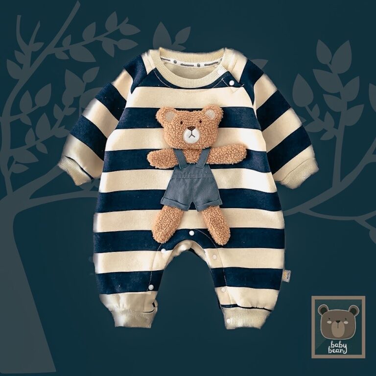 Baby full body romper in navy and cream stripe has been placed on a dark, grey blue background. Laid down flat to show style of the bodysuit which is thick and closes with metal fasteners at the bottom. Huge teddy bear wearing dungarees has been sewn to the front of the whole bodysuit romper.