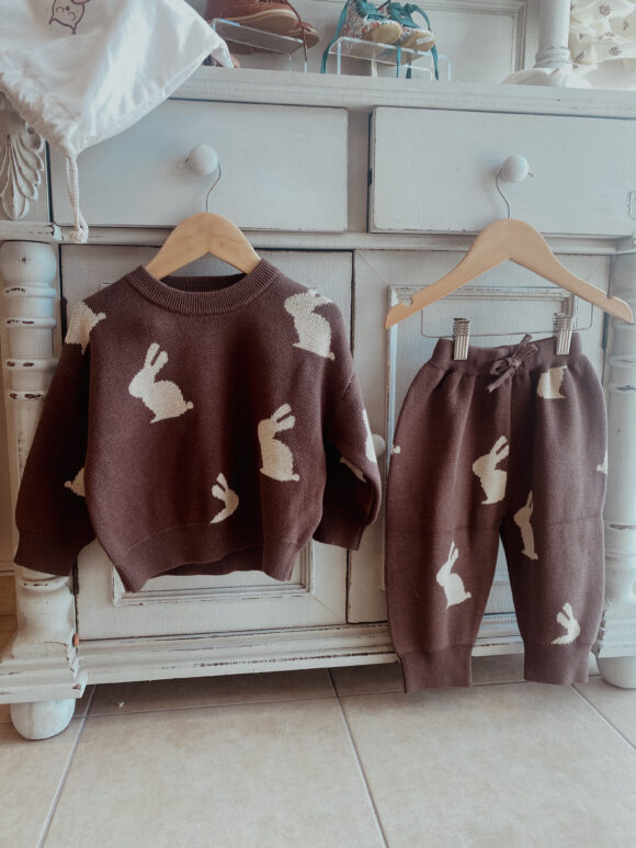 This image shows a brown knitted toddler clothing set. The set depicts a jumper and a matching pair of jogging bottoms with drawstring WAIST. the fabric is cotton and it is brown with cream bunny rabbits printed on it.