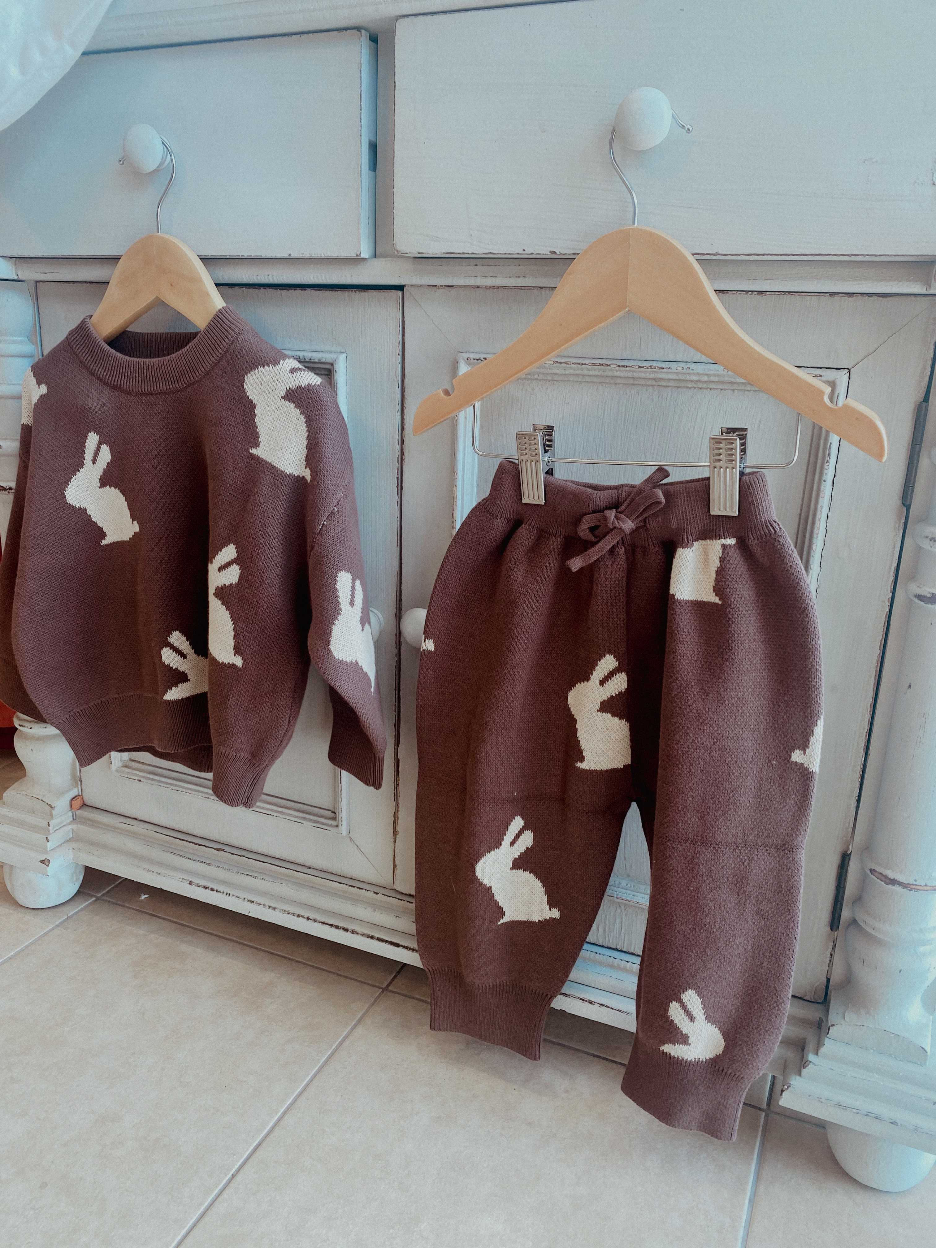 a children's knitted set of jumper and pants is hanging from wooden hangers. The set is knitted jacquard and is brown with cream bunny print on it. The jumper and jogging pants with tie-string waist are hanging from the drawers of a vintage piece of furniture.