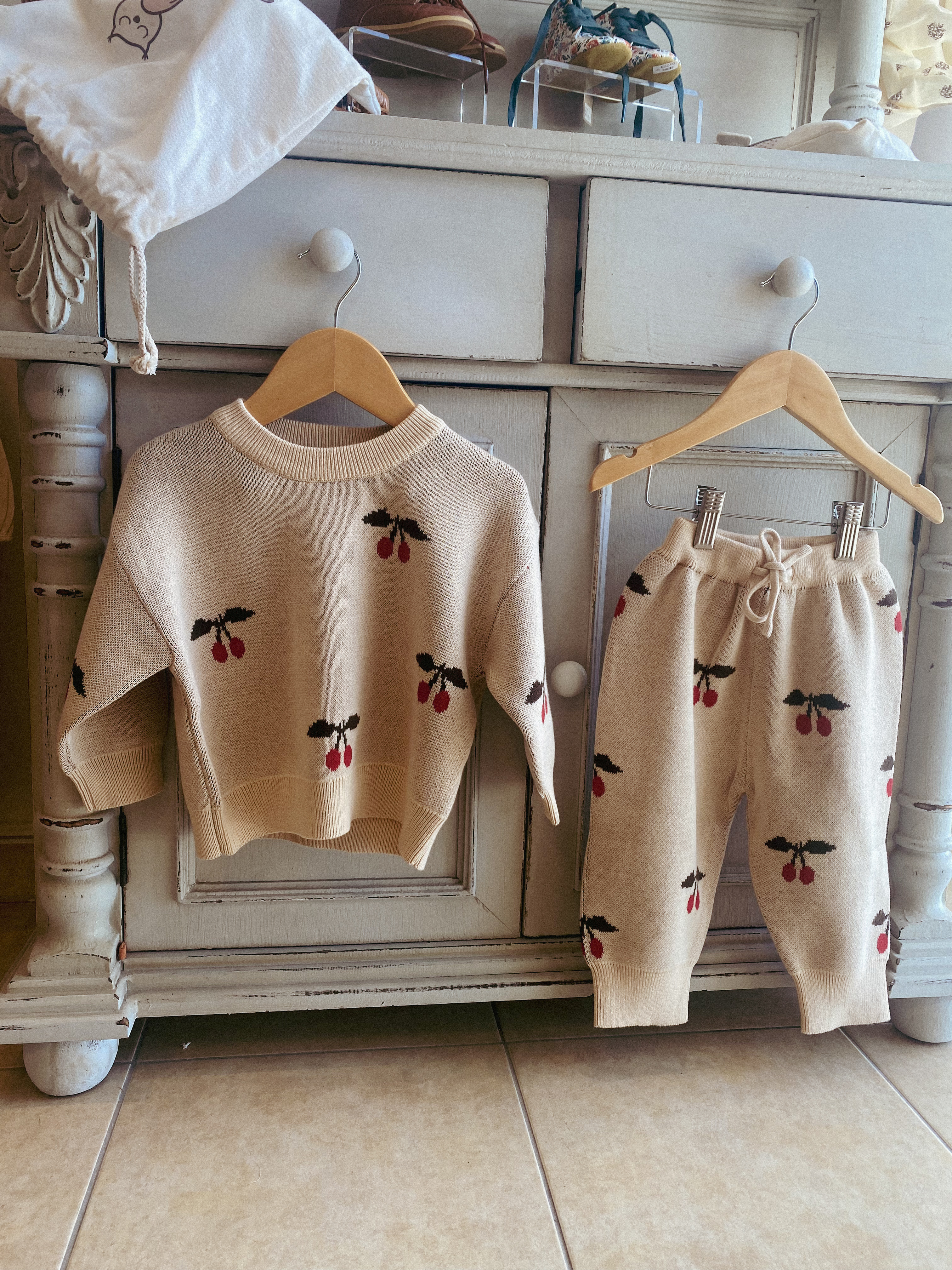 creamy white cotton jacquard knitted toddler sweatshirt and matching bottoms set is hanging from a vintage piece of furniture. the pants are hanging next to the top and the image shows how the clothes are cut and the style they have also the pattern which is red and brown cherries.
