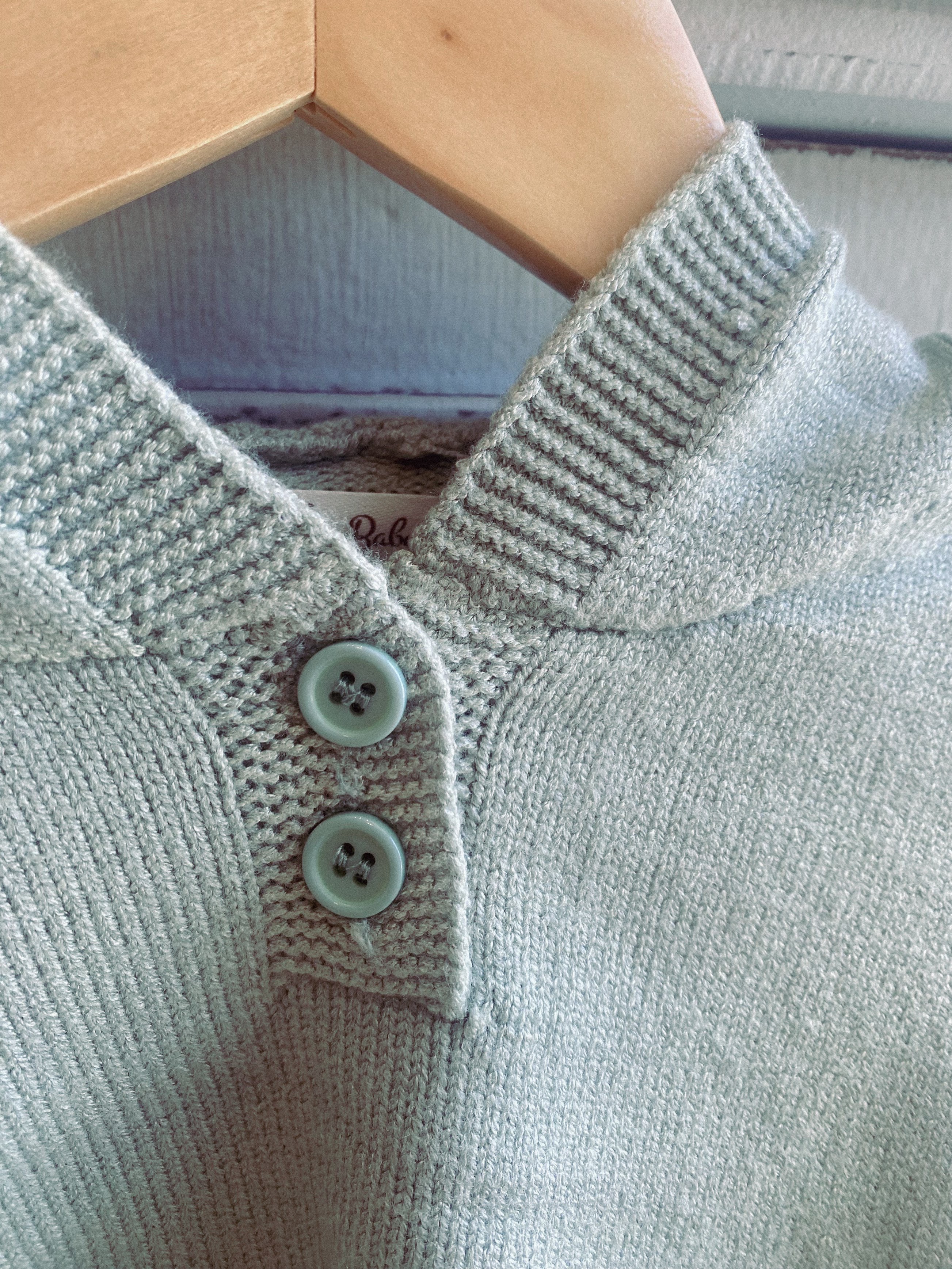 Close up picture of a kids knitted set. the picture depicts the top part of the knitted set and is the colour sage. the top part shows two buttons and the hood of the jumper.