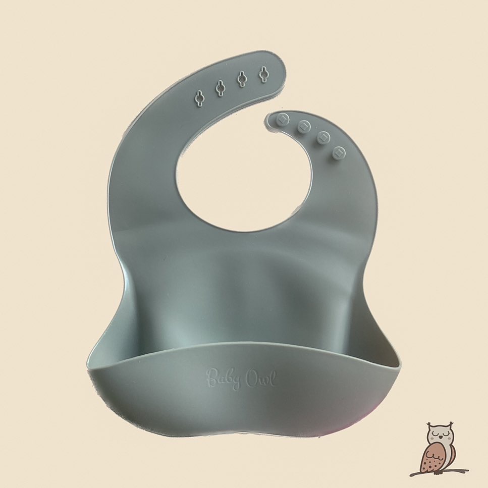 silicone bib with brand name baby owl printed on front. the bib has a piece at the bottom to collect food that falls. the colour is sage.