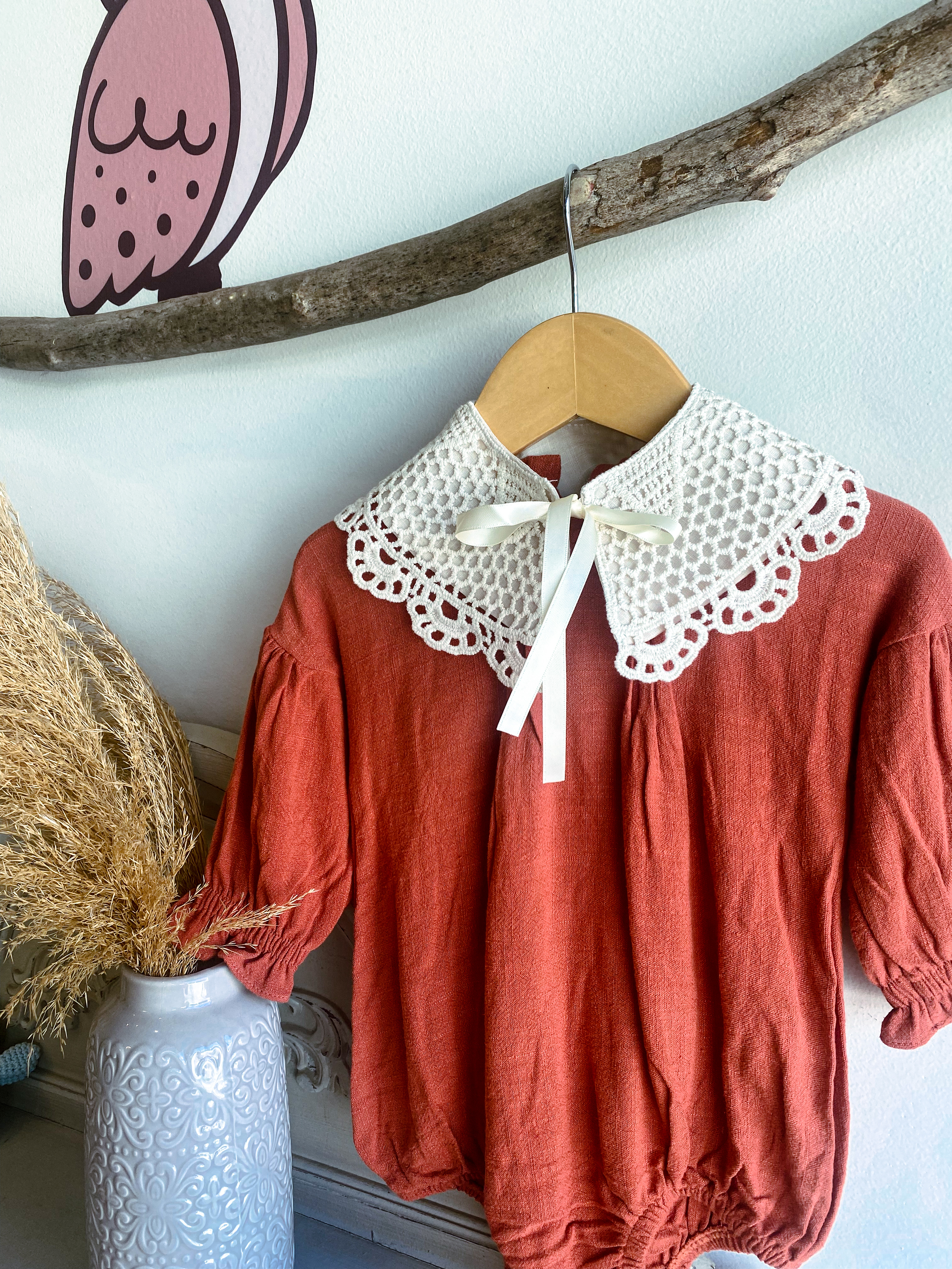 A red linen baby romper with flounced sleeves is displayed with a vintage lace bib accessory. The bib is cream and ties at the front with a ribbon.