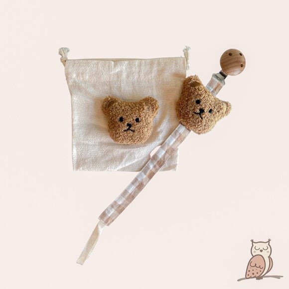 cotton pacifier clip with gingham fabric strip. sewn to the top of the fabric strip is a fluffy teddy bear head. the same teddy head is sewn onto the linen bag that comes with the pacifier clip for trendy and safe keeping.