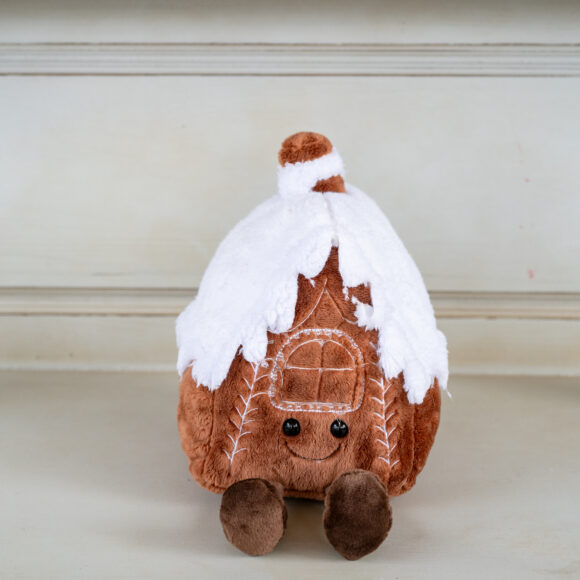 Christmas teddy bear photographed on a vintage piece of furniture. The gingerbread house is brown with a cute face and hanging feet and a white cotton roof depicting snow falling from the roof.