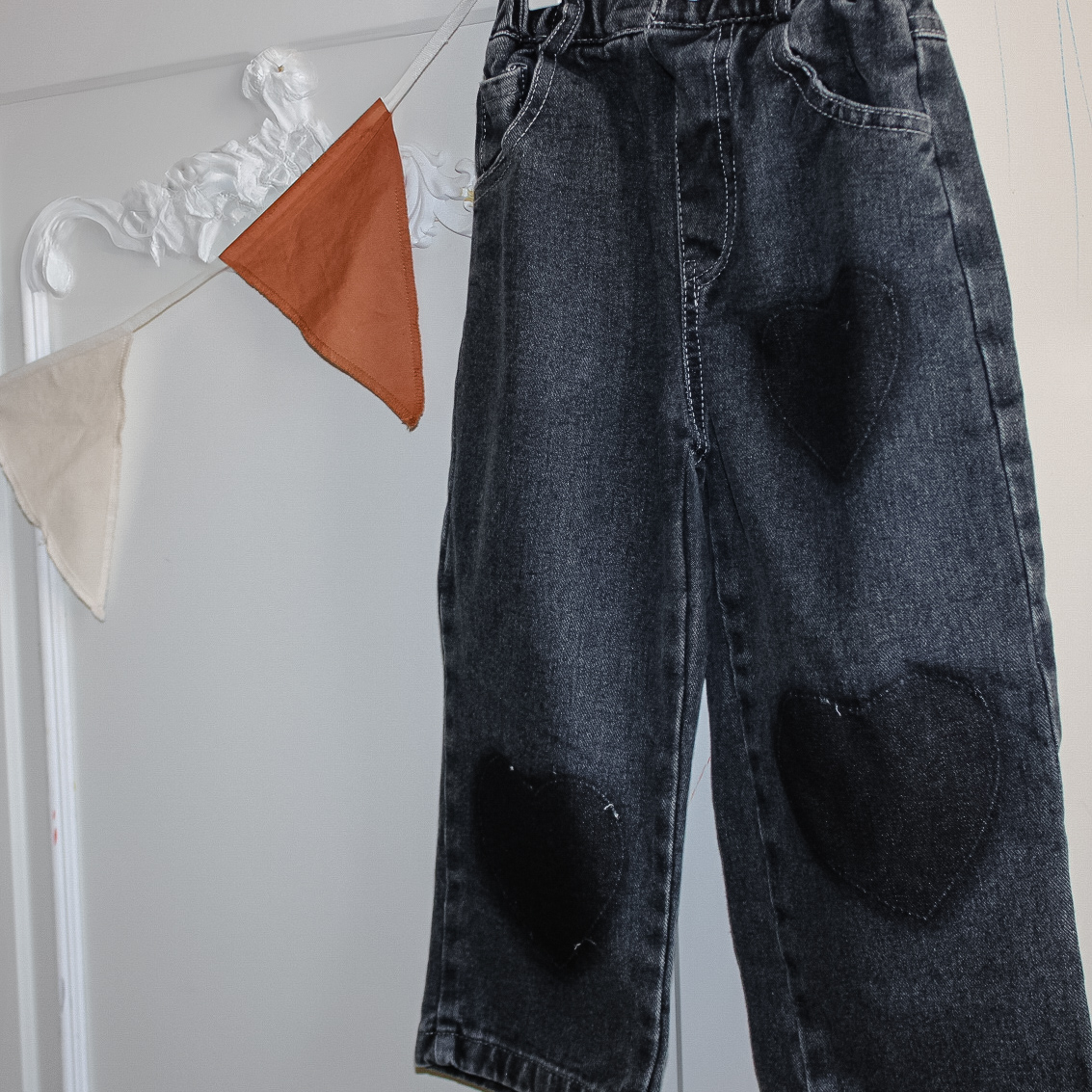 Pair of girls black jeans hanging from a vintage wardrobe. The jeans are in a distressed black with three darker hearts, two on one leg and one on the other.